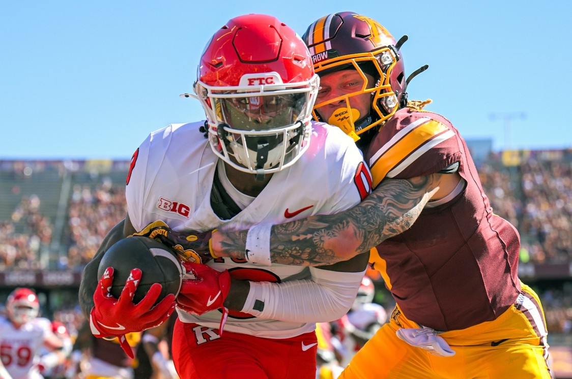 American football action: a ball carrier in white and red grips the ball while a defender in maroon and gold wraps him up during a sunny game.